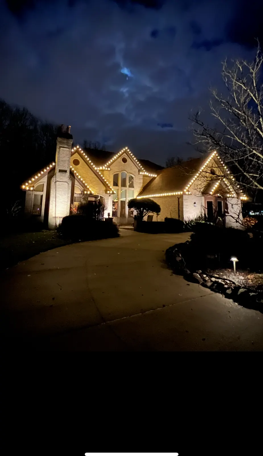 Light stone two-story home at night with warm white Christmas lights on every gable, a lit chimney, and an arched center window