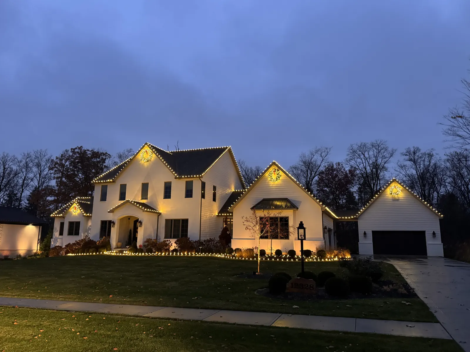 White two-story home at twilight with four matching lit Christmas wreaths centered on the gables and warm white C9 lights on every peak